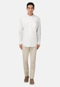 Camicia bianca con bottoni, taschino sul petto, maniche lunghe e orlo arrotondato, abbinata a pantaloni beige e scarpe marroni. Finitura in tessuto liscia.