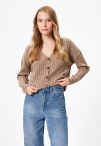 Cardigan marrón con canalé, escote en V y cuatro botones oscuros. Lleva puesto sobre unos vaqueros de cintura alta de color azul claro. Manga larga y corte relajado.