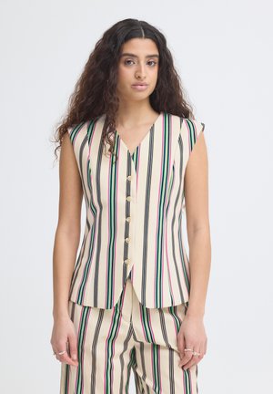Kvinde med langt krøllet hår iført en ærmeløs beige top og bukser med lodrette sorte, grønne og lyserøde striber samt knapper foran.