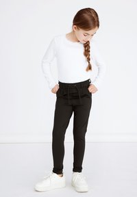 Zwarte tappered broek met een touwsluiting in de taille, gecombineerd met een witte langslijfshemden en witte sneakers. Het ontwerp heeft voorzakken.