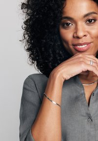 Szara lniana koszula z kołnierzykiem, srebrna bransoletka i smukły srebrny naszyjnik. Kręcone ciemne włosy oprawiają twarz. Gładka skóra z delikatnym makijażem.