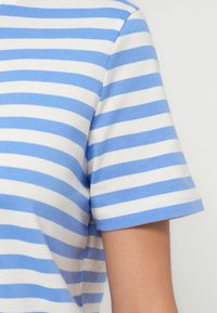 Sinine-valge horisontaalse triibuga lühikeste varrukatega särk inimesel, näidates õlavart ja õlga ühevärvilise tausta ees.