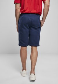 Navyblauwe shorts van zacht materiaal met een elastische tailleband, die tot halverwege de dij komt. Gecombineerd met witte sneakers, gezien vanuit de achterkant.
