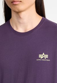 Lila pamut póló kerek nyakkal. A bal mellrészen egy kis, világos színű "Alpha Industries" logó található.