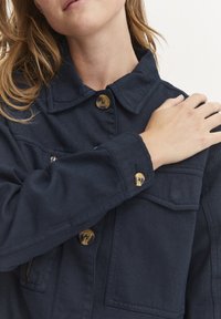 Marineblå denimjakke med spiss krage, to brystlommer og messingknapper; har en strukturert stoff og en avslappet passform.