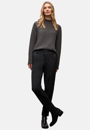 Grauer Strickpullover mit hohem Kragen, kombiniert mit schwarzen Hosen mit glatter Textur und schlanker Passform. Modell trägt schwarze Stiefeletten.