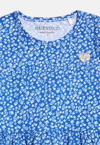 Μπλε φόρεμα με λευκό λουλουδάτο μοτίβο, στρογγυλή λαιμόκοψη, λογότυπο GUESS μέσα στον γιακά και χρυσό έμβλημα GUESS σε σχήμα καρδιάς στο στήθος.