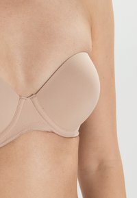 Beige, stroppeløs BH med glatte, polstrede kopper, som har vertikale sømmer og en subtil glans, designet for en sømløs passform under klær.