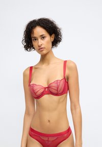 Sutiã vermelho com aros e copas de malha transparente, apresentando detalhes bordados. Acompanhado de uma tanga vermelha transparente correspondente. Ambos têm alças finas.