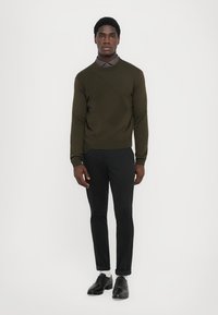 Maglione lavorato a maglia verde oliva con un motivo diagonale, abbinato a pantaloni neri slim-fit e scarpe nere, creando un aspetto stratificato.