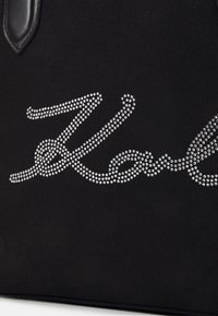 Black canvas tote bag featuring a silver rhinestone signature design. Smooth black leather accents complement the textured material.