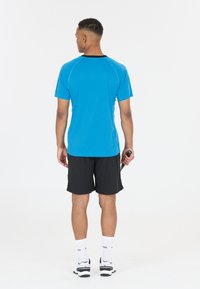 Blå sportströja med korta ärmar och svarta detaljer, i kombination med svarta shorts och vita sportskor. Modellen håller en racket.