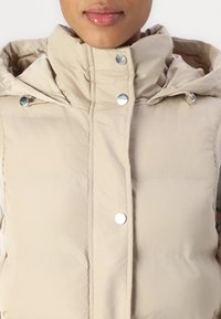 Gros plan sur une personne portant un gilet matelassé beige avec des boutons-pression en métal et une capuche sur un fond uni.