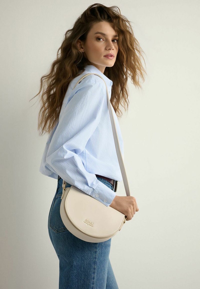 Kobieta o długich, falowanych włosach, ubrana w jasnoniebieską koszulę w paski i dżinsy, trzymająca jasnobeżową torebkę na ramię w kształcie półksiężyca.