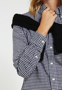 Checkered shirt in navy and white with buttons, featuring long sleeves and a relaxed fit; black knitwear draped over shoulders.