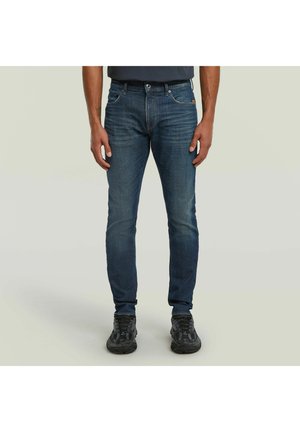 Mann trägt eng anliegende dunkelblaue Jeans und schwarze Sportschuhe, steht vor einem schlichten hellen Hintergrund.