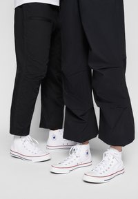 Pantalon noir ample à devant plat avec poches, associé à des baskets montantes blanches arborant un logo étoile bleu et des accents rouges.