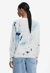 Weißer Pullover mit blauem Batikmuster, langen Ärmeln und gerippten Bündchen. Kombiniert mit hellblauen, locker sitzenden Jeans.