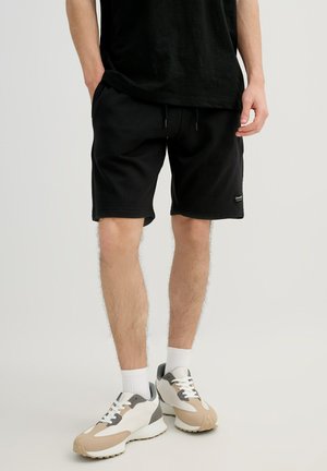 Hombre con pantalones cortos negros, calcetines blancos y zapatillas beige, blancas y grises, de pie contra un fondo claro y liso.