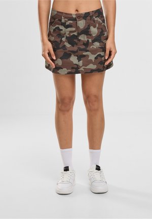 Naine, kes kannab pruuni ja rohelise kamuflaažiga miniseelikut, valgeid crew-sokke ja valgeid tenniseid, seisab ühevärvilise hele tausta ees.