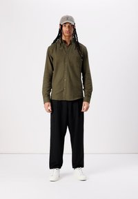 Camisa verde aceituna de botones con bolsillo frontal, pantalones negros de corte holgado y zapatillas blancas. Una gorra gris completa el conjunto.