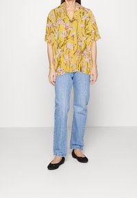 Kurzärmeliges gelbes Hemd mit pinkem Blumenmuster, kombiniert mit hellblauen geraden Jeans und schwarzen flachen Schuhen.