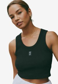 Crni rebrasti crop top s okruglim dekolteom, na kojem se nalazi tekst "se lf." u bijeloj boji. Tkanina izgleda mekano i ima uski kroj.