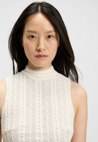 Młoda kobieta z długimi ciemnymi włosami, ubrana w białą koronkową bluzkę bez rękawów o wysokim dekolcie, na jednolitym jasnym tle.