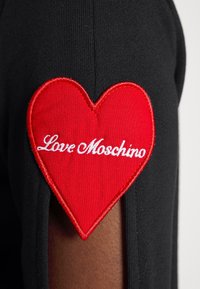 Patch en forme de cœur rouge avec le texte blanc « Love Moschino » cousu sur une manche en tissu noir.