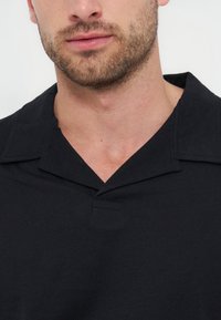 Άνδρας με γένια, που φοράει μαύρο πουκάμισο με γιακά και V-λαιμόκοψη, μπροστά από απλό ανοιχτό φόντο.