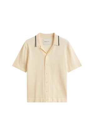 Chemise beige à manches courtes avec boutons et col présentant deux bandes noires, posée à plat avec l'étiquette de la marque visible à l'intérieur du col.