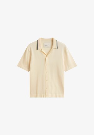 Beige overhemd met korte mouwen en knopen, voorzien van een kraag met zwarte dubbele strepen, plat neergelegd met zichtbaar merkembleem aan de binnenkant van de kraag.