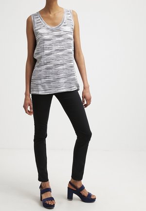 Personne portant un haut sans manches blanc à rayures noires, un pantalon slim noir et des sandales plateformes bleu marine, debout sur un fond blanc.