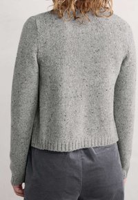 Osoba oblečená v šedém melírovaném pleteném svetru a tmavě šedých kalhotách, zachycena zezadu na jednotném světlém pozadí.