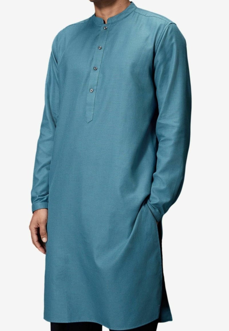 Man in een lichtblauw, textuur gekleed kurta met lange mouwen, een met knopen gesloten mandarin kraag en een hand in zijn zak.