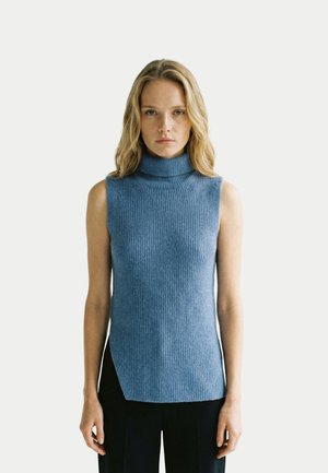 Femme portant un pull à col roulé côtelé bleu sans manches et un pantalon noir, debout devant un fond clair uni, face à l'avant.
