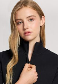 Giacchino nero con zip e colletto alto, realizzato in un tessuto testurizzato. Il modello tiene la zip con una mano, mostrando un primo piano.