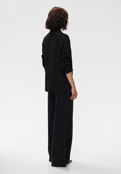 Personne aux cheveux courts et bouclés, vue de dos, portant une chemise noire à manches longues et un pantalon noir à jambes larges sur un fond uni.