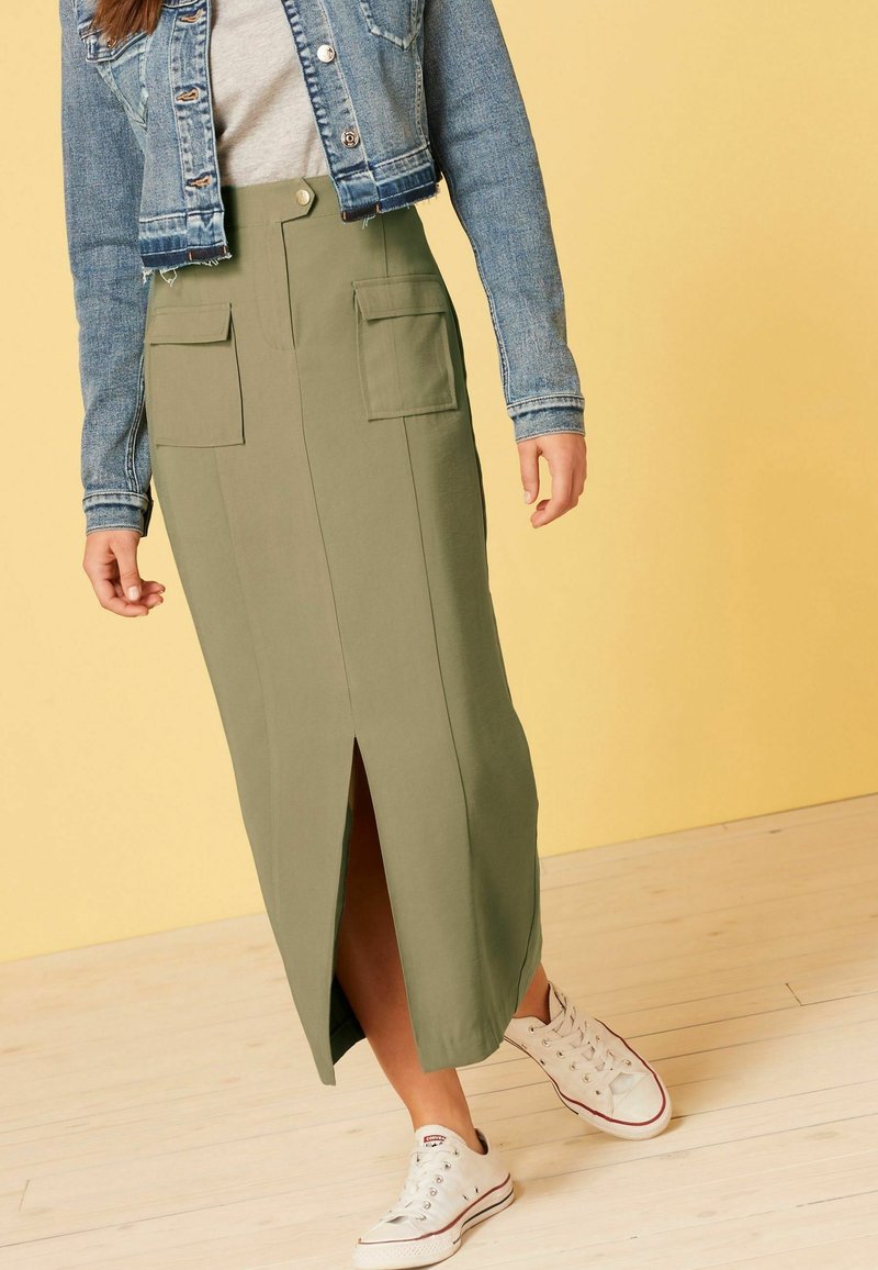 Falda maxi verde oliva con una abertura en la parte delantera, dos grandes bolsillos frontales y cierre de botón. Llevada con una blusa gris y una chaqueta de mezclilla.