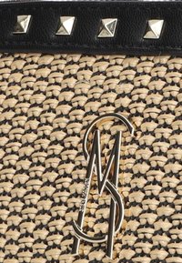 Woven natural fiber bag with a black textured leather strap featuring silver pyramid studs. Prominent metal logo in gold.