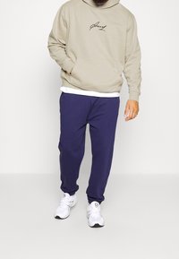 Hoodie beige con logo nero, tasca frontale; abbinato a pantaloni jogger blu navy. Sneakers bianche con accenti neri e suola dettagliata.