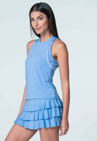 Mujer con cabello largo y oscuro, vestida con una blusa sin mangas de color azul claro y una falda de volantes en capas a juego, contra un fondo claro y sencillo.