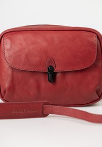 Red leather bag with a smooth texture, rectangular shape, fold-over flap, and a black clasp. Includes an adjustable strap embossed with the brand name.