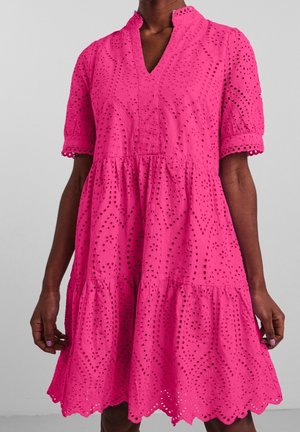 Vestido de color rosa brillante hasta la rodilla con patrones de ojales, mangas cortas, escote en V y falda con volantes en capas, en una persona de piel oscura.