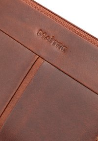 Brown leather pouch with a zip closure. Features embossed "MAÎTRE" logo and a textured surface with a smooth finish and distinct stitching.