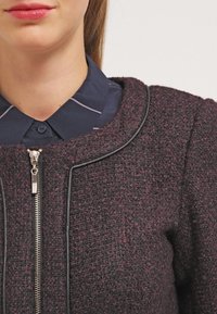 Vínová texturovaná bunda s kulatým výstřihem, černým lemováním a stříbrným zipem, nošená přes tmavě modrou košili s límečkem.