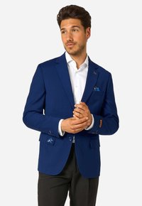 Vīrietis pielāgo zilo žaketi virs baltās krekls un melnām biksēm, ar rakstainu kabatas pledu un smalku izšuvumu uz piedurknes un kabatas atloka.