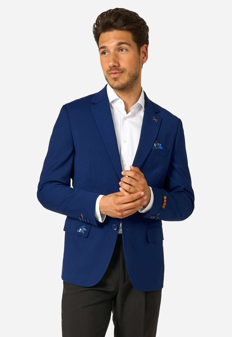 Vīrietis pielāgo zilo žaketi virs baltās krekls un melnām biksēm, ar rakstainu kabatas pledu un smalku izšuvumu uz piedurknes un kabatas atloka.