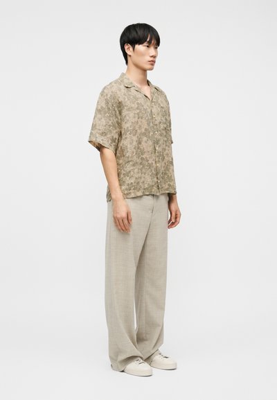 Άνδρας που στέκεται με μπεζ κοντομάνικο πουκάμισο με floral σχέδιο, ανοιχτόχρωμο παντελόνι με φαρδιές μήνες και λευκά αθλητικά παπούτσια, μπροστά σε απλό λευκό φόντο.