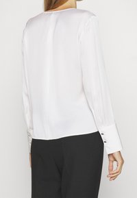 Bílá hedvábná blůza s dlouhými rukávy, kulatým výstřihem a knoflíkovými manžetami; jemný lesk a hladká textura; kombinováno s černými kalhotami.